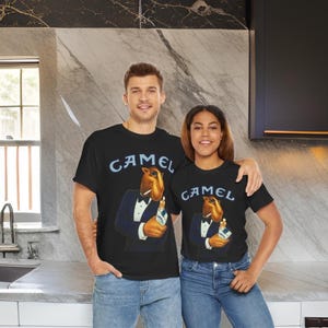 May include: Two people wearing black t-shirts with a vintage Camel cigarette ad featuring a cartoon camel in a tuxedo. The ad text reads "Camel".