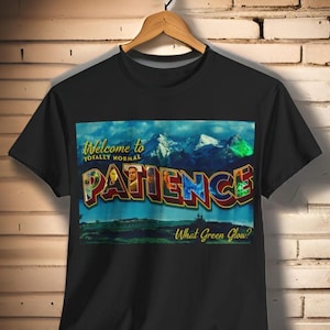May include: Black t-shirt featuring a vintage postcard design with a mountain scene and the text "Welcome to Totally Normal Patience. What Green Glow?"