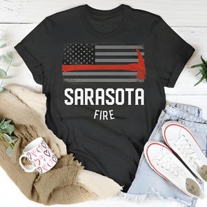 May include: Black t-shirt featuring a red, white and blue American flag and an axe graphic. The text "Sarasota Fire" is printed below the flag.