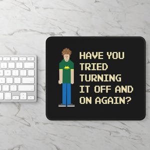 May include: Black rectangular mousepad with the text "HAVE YOU TRIED TURNING IT OFF AND ON AGAIN?" and a pixelated graphic of a person. A white keyboard is visible in the background.