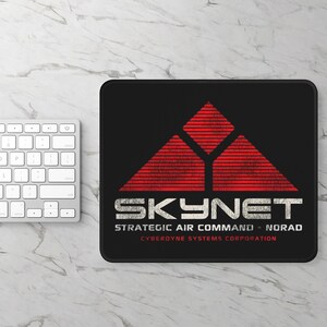 May include: Black mouse pad with a red and white Skynet logo. The logo features a stylized triangle with the text "Skynet" in white and the words "Strategic Air Command NORAD" and "Cyberdyne Systems Corporation" in white below the logo.