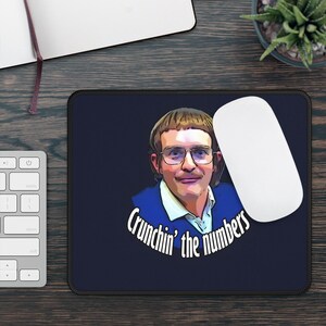 May include: A navy blue mousepad featuring a cartoon portrait of a man wearing glasses and a blue blazer. The text "Crunchin' the numbers" is written in a white, curved font below the portrait. A white computer mouse is on the right.