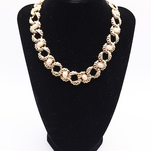 May include: A gold-toned necklace with a repeating floral design and oval, light pink accents. The necklace is displayed on a black velvet bust, showcasing its intricate details and elegant style. The chain links are connected by a series of floral-shaped links.