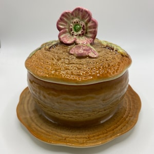 May include: A ceramic butter dish with a textured, honey-brown finish resembling a woven basket. The lid features a sculpted, three-dimensional pink flower with green leaves on top. The base is a matching brown plate with a scalloped, wavy edge.