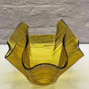 May include: An amber-coloured glass bowl with a distinctive folded design. The bowl features a textured surface with horizontal lines and a dark rim. The folded edges create a wave-like appearance, adding visual interest to the piece.