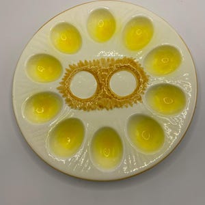 May include: A cream-coloured ceramic escargot plate with eleven indentations for snails. The plate has a gold rim and a central design with two circular spaces and a textured, sunburst-like pattern.
