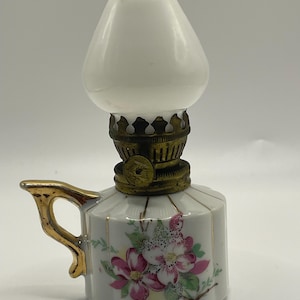 May include: A miniature oil lamp featuring a white glass shade and a floral pattern. The ceramic base is white with pink and white flowers, gold detailing, and a gold handle. The metal parts have a brass finish.