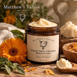 Könnte beinhalten: Ein bernsteinfarbenes Glas mit cremefarbenem Inhalt und einem beigen Etikett, das eine Kuh-Illustration sowie den Text Matthew's Tallow, Organic Tallow Calendula Mineral Cream, Pure & All Natural, Net wt. 227 g zeigt. Umgeben von orangefarbenen Blüten.