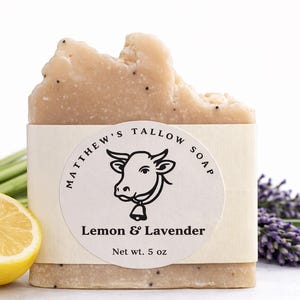 May include: A bar of Matthew's Tallow Soap, featuring a Lemon & Lavender scent, with a cow logo on the label. The soap is light brown with dark specks. A sliced yellow lemon, green stems, and purple lavender sprigs are also visible.