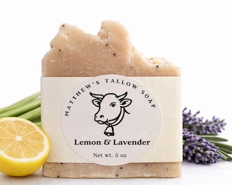 May include: A rectangular bar of beige tallow soap with visible dark specks, wrapped in a paper label reading Matthew's Tallow Soap Lemon & Lavender Net wt. 5 oz with a cow illustration, positioned next to a yellow lemon slice and lavender sprigs.