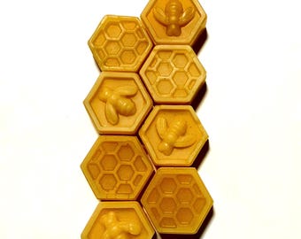 May include: A collection of eight hexagonal, yellow-orange soap bars arranged vertically. Each bar features a honeycomb pattern or a bee design. The soaps are on a white background, creating a clean and simple aesthetic.