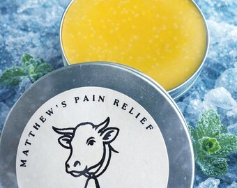 May include: A silver tin of Glacier Balm with a yellow, textured balm inside. The lid features a cow illustration and the text "Matthew's Pain Relief." The net weight is 2 oz. The balm is set on a bed of ice.