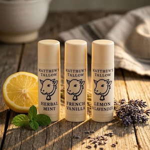 K&ouml;nnte beinhalten: Drei Lippenbalsam-Tuben mit der Aufschrift "Matthew's Tallow" und einem Kuh-Logo, in den Geschmacksrichtungen Herbal Mint, French Vanilla und Lemon & Lavender. Eine Zitronenh&auml;lfte, Minze und Lavendelzweige sind ebenfalls zu sehen.
