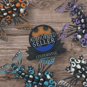 May include: A black and blue round badge with the text "AUTHORIZED SELLER" in white and the text "CINDERRWING!" in white on a black ribbon. The badge is surrounded by four colorful 3D printed butterfly figurines.