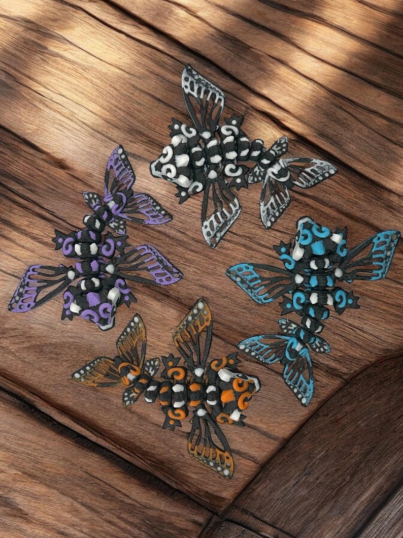 May include: Four decorative butterflies made of black, white, purple, blue, and orange beads. The butterflies are arranged on a wooden surface.