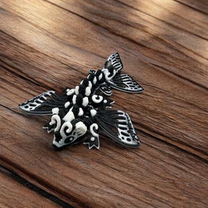 May include: A black and white 3D printed figurine of a fish with intricate details. The fish has large wings and a detailed face.