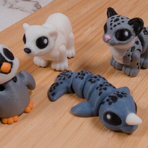 May include: Four small, colorful animal figurines on a wooden surface. The figurines include a penguin, a white bear, a gray leopard with black spots, and a blue rhino-like creature with a white horn. The penguin has orange feet.