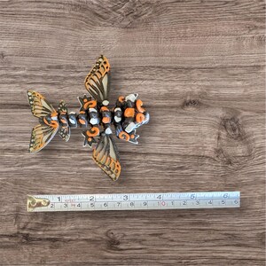 May include: A black, orange, and white 3D printed fish with butterfly wings. The fish is approximately 4 inches long and is sitting on a wooden surface.