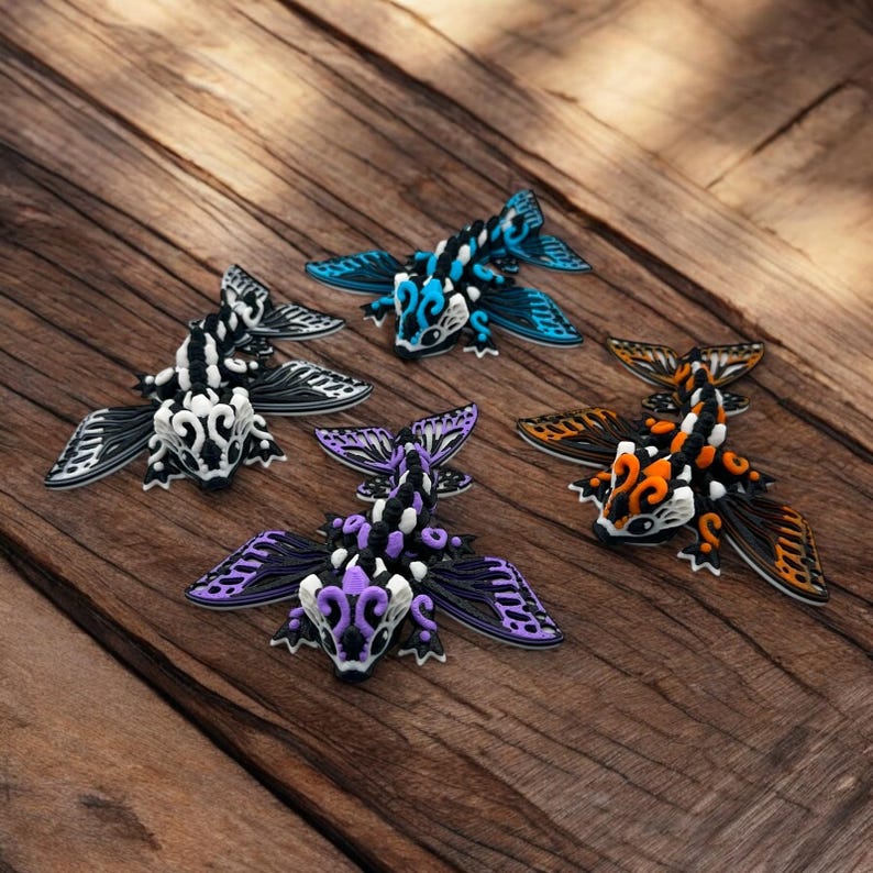 May include: Four colorful, intricately designed, 3D printed dragonflies with black, white, and various colored accents. The dragonflies are arranged on a wooden surface.