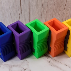 May include: Five brightly coloured pencil sharpeners in blue, purple, green, orange, and yellow. Each has a rectangular top and a ridged, curved body. The sharpeners are made of plastic and sit on a marble surface.