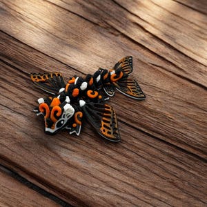 May include: A black, orange, and white decorative figurine of a stylized fish with wings. The figurine is made of a textured material and has intricate details.