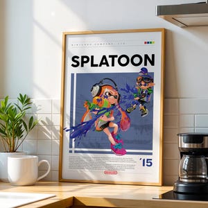 Puede incluir: Póster enmarcado del videojuego "Splatoon". La obra de arte muestra a dos personajes en acción, con colores vibrantes y salpicaduras de tinta. El póster tiene un marco de madera y se exhibe en una encimera de cocina.