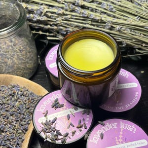 May include: A close-up of several jars of lavender-infused herbal balm. The jars are dark amber glass, with a light yellow balm inside. The labels read "lavender lush" and "hand crafted organic herbal balm". Dried lavender sprigs and loose buds are scattered around the jars.