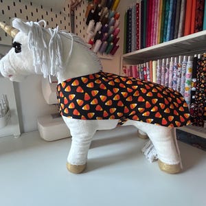Puede incluir: Un unicornio de peluche blanco con cuerno y pezuñas doradas, que lleva una capa negra estampada con caramelos de maíz naranja, amarillo y blanco. El unicornio tiene una melena y cola blancas. El fondo incluye suministros de costura y rollos de tela.