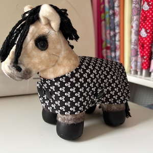 Op de afbeelding: Een pluchen paard met een beige lichaam, zwarte manen en zwarte hoeven. Het draagt een zwarte jas met witte doodshoofden. Het speelgoed heeft grote, donkere ogen en staat op een wit oppervlak.