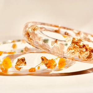 May include: Two clear resin bangles with embedded dried flowers and gold leaf. One bangle is resting on top of the other. The bangles have a transparent appearance, showcasing the natural elements within.