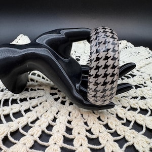 May include: A thick, rounded bangle bracelet featuring a classic black and white houndstooth pattern. The surface of the bracelet has a smooth, glossy finish and includes a small, rectangular embedded detail with a grid of tiny silver-toned studs.