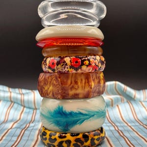 May include: A vertical stack of eight assorted bangles. The collection includes a clear bangle, a solid cream bangle, a translucent red bangle, a brown marbled bangle, a floral print bangle, a faceted brown wood-grain bangle, a cream bangle with a blue feather print, and a yellow and black leopard print bangle.