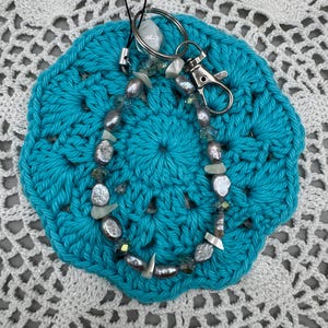 Può includere: Un ciondolo per telefono con perle grigie, piccoli cristalli iridescenti e frammenti di conchiglie bianche. L'accessorio include un moschettone in metallo color argento e un cordino nero per il fissaggio al dispositivo.