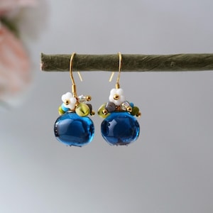 Può includere: Orecchini pendenti con una perla di vetro sferica blu scuro. Ogni orecchino ha un gancio color oro ed è decorato con piccole perline bianche a forma di fiore e altre perline colorate. Gli orecchini sono esposti su uno sfondo morbido e sfocato.