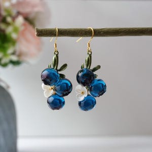 May include: A pair of dangle earrings featuring clusters of dark blue, translucent orbs resembling blueberries. Each cluster includes small white flower accents and green leaves, suspended from gold-colored hooks.