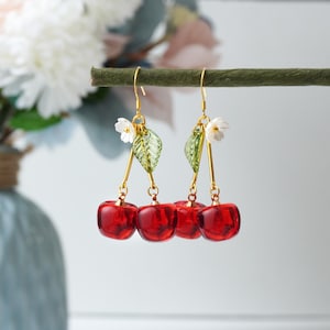 May include: Dangle earrings featuring clusters of red, translucent cherries. The earrings have gold-colored hooks and accents, with small white flowers and green leaves. The earrings are displayed against a soft, blurred background.