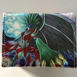 Può includere: Un portafoglio con una colorata illustrazione di una creatura fantasy. Il design presenta una figura alata con dettagli rossi, verdi e neri, su uno sfondo blu e viola. Il portafoglio ha un bordo cucito.