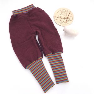 Könnte beinhalten: Bordeauxfarbene Babyhose mit Rippstruktur. Die Bündchen und der Bund weisen farbenfrohe horizontale Streifen auf. Die Hose wird auf einer weißen Oberfläche präsentiert. Eine Holzscheibe mit Text und eine kleine weiße Schaf-Figur befinden sich im Hintergrund.