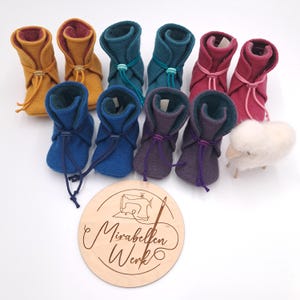 Könnte beinhalten: Eine Auswahl an Baby-Booties in Gelb, Türkis, Magenta, Blau und Lila. Jeder Bootie hat einen Kordelzugverschluss. Eine Holzscheibe mit dem Text "Mirabellen Werk" ist ebenfalls vorhanden, zusammen mit einem kleinen Wollschaf.