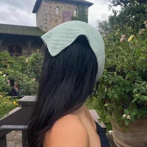 May include: A light green crochet head covering. The head covering is worn over dark hair. The background features a stone building with a green roof and lush greenery. The head covering is a stylish accessory.