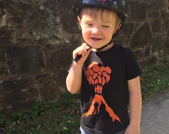 May include: A young child wearing a black helmet with the word "Wipeout" on it, a black t-shirt with an orange volcano design, and blue denim shorts.