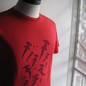 May include: Red t-shirt featuring a graphic design of several hammerhead sharks in a dark red colour.