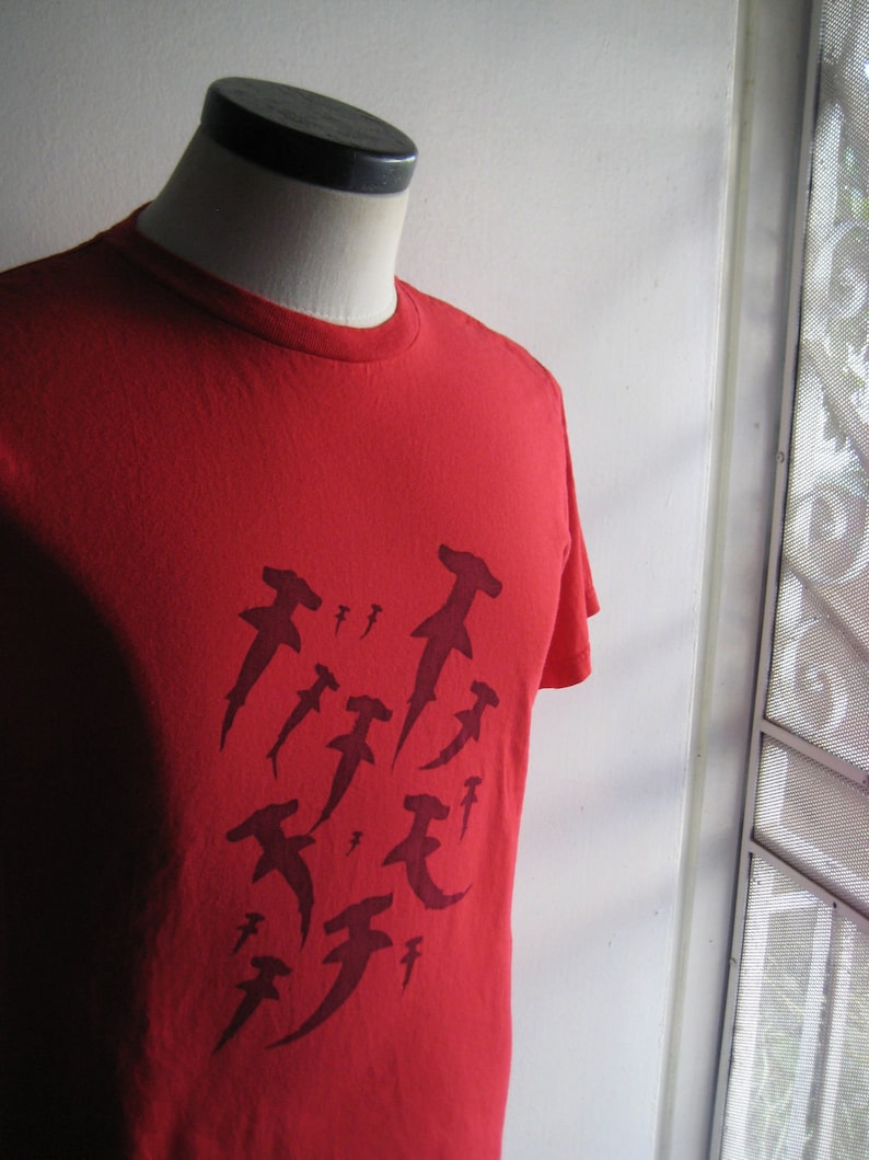 May include: Red t-shirt featuring a graphic design of several hammerhead sharks in a dark red colour.