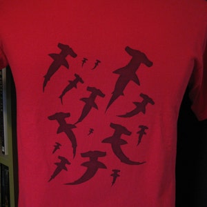 May include: Red t-shirt featuring a black silhouette design of several hammerhead sharks.