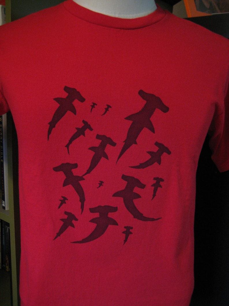May include: Red t-shirt featuring a black silhouette design of several hammerhead sharks.