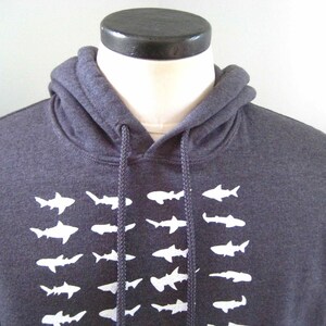 Op de afbeelding: Een donkergrijze hoodie met een witte grafische print van een school haaien. De hoodie heeft een trekkoord.