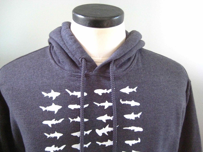Op de afbeelding: Een donkergrijze hoodie met een witte grafische print van een school haaien. De hoodie heeft een trekkoord.