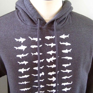 Op de afbeelding: Een donkergrijs hoodie met een witte grafische print van een rij haaien. De haaien kijken allemaal dezelfde kant op en zijn verticaal gerangschikt.