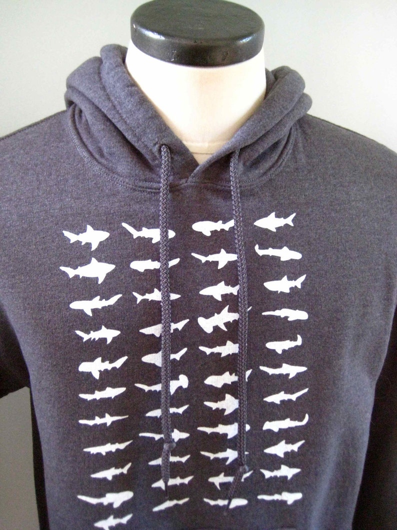 Op de afbeelding: Een donkergrijs hoodie met een witte grafische print van een rij haaien. De haaien kijken allemaal dezelfde kant op en zijn verticaal gerangschikt.