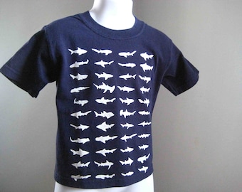May include: A navy blue t-shirt with a white shark silhouette pattern. The shirt has short sleeves and a crew neck.